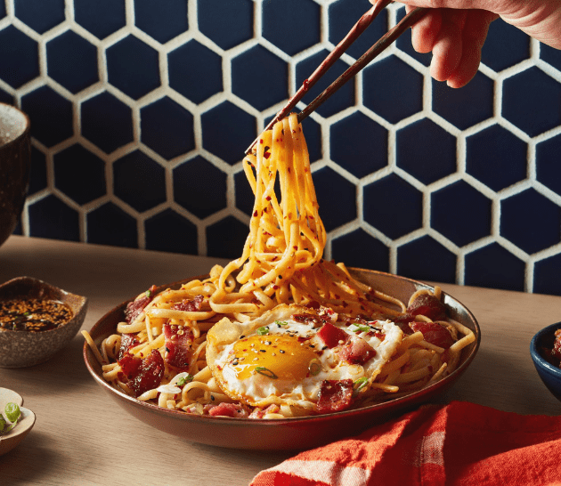 Bacon chili oil noodles in a brown bowl being picked up with chopsticks